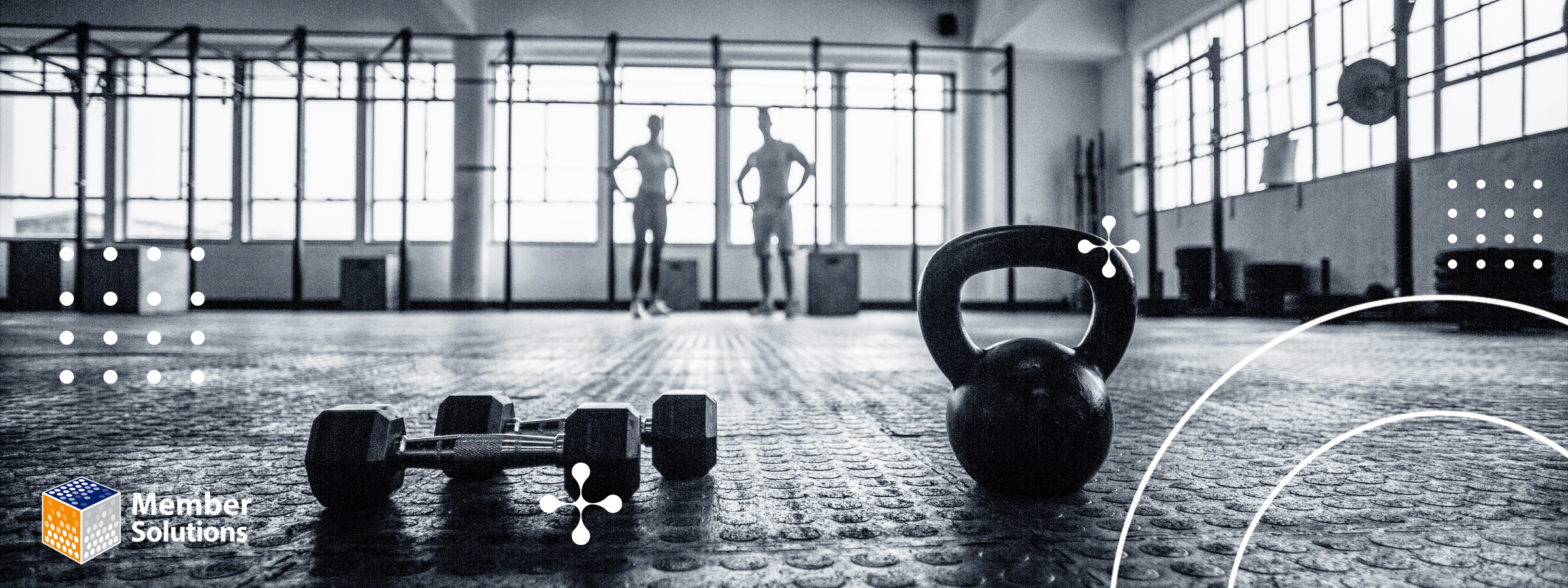 In a dimly lit gym, the silhouettes of two people appear in the background, while a kettlebell and dumbbells are keep in the foreground