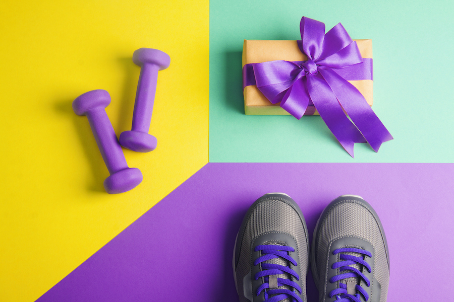 flat lay composition with gray shoes, purple dumbbells and craft gift box with violet bow on yellow purple and cyan paper background.