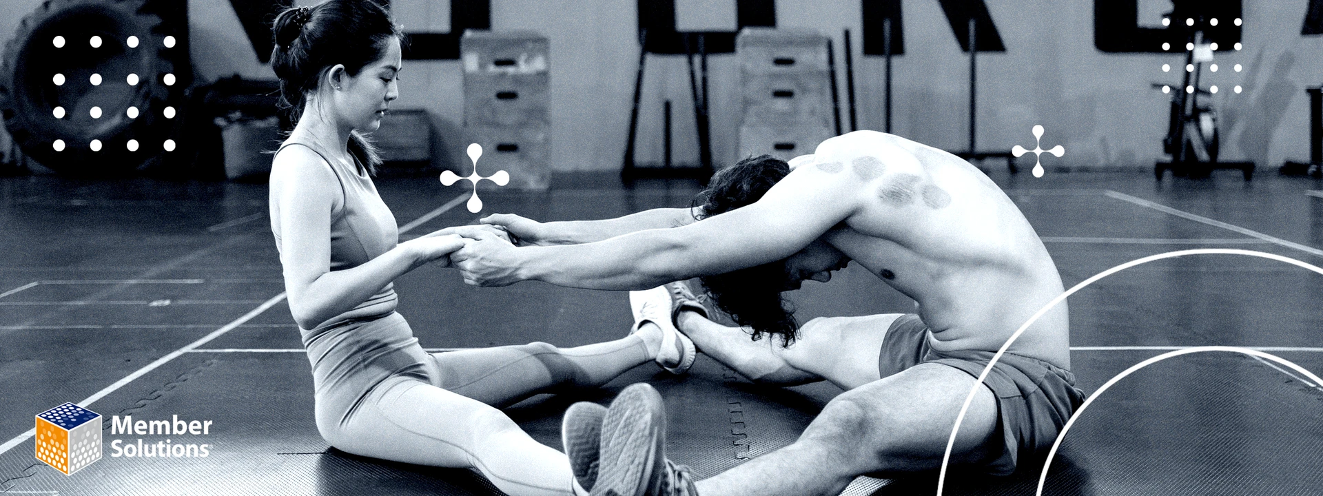 Two people stretch on an industrial-style gym floor, holding hands for support, with large tires and fitness equipment in the background