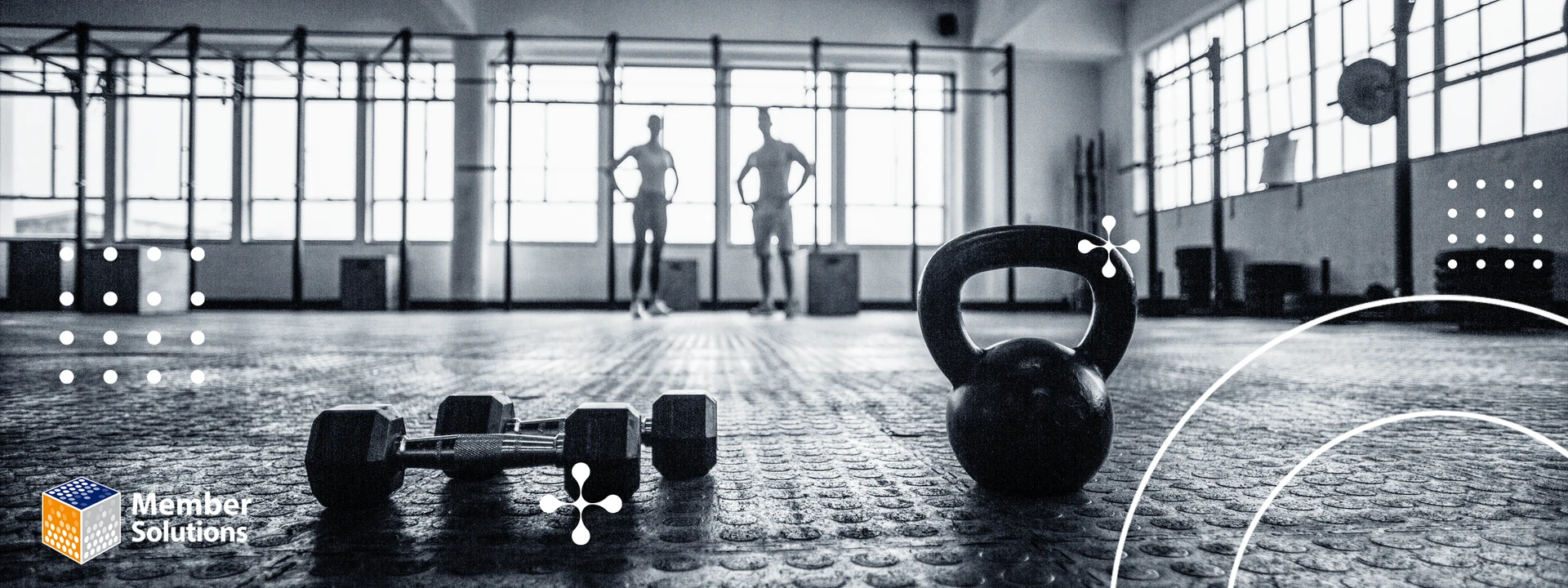 In a dimly lit gym, the silhouettes of two people appear in the background, while a kettlebell and dumbbells are keep in the foreground