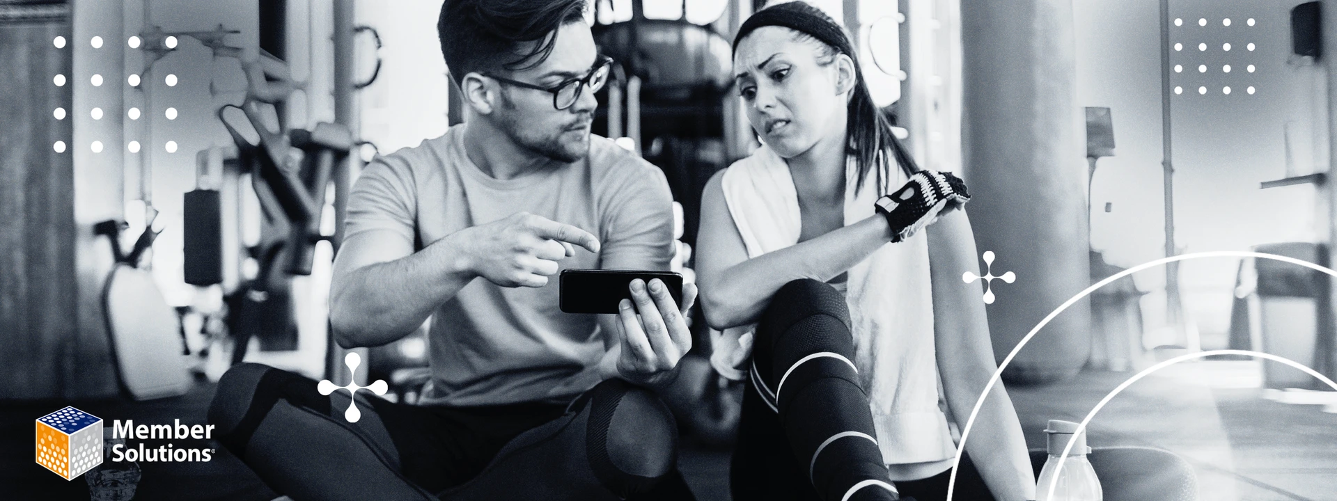 A man and woman in workout attire sit on a gym floor. The man points at a smartphone, and the woman appears pensive. Gym equipment is in the background