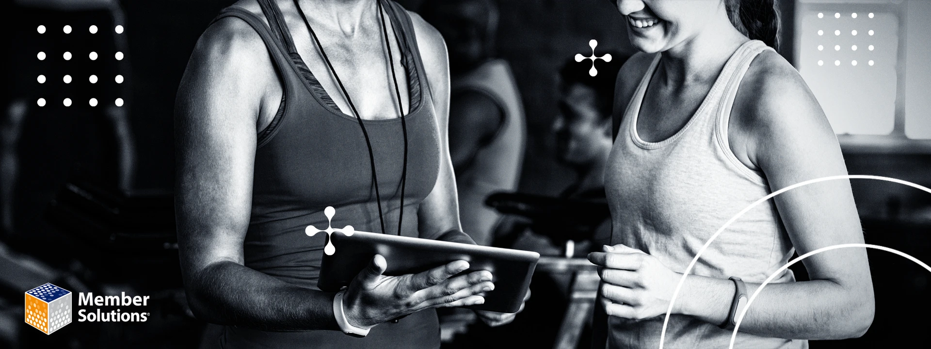 Two people in athletic clothing are smiling and looking at a tablet in a gym