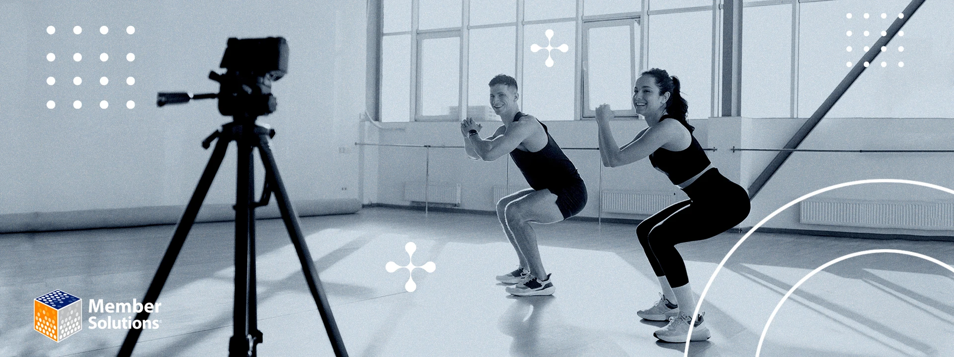 Two people exercise in a gym, performing squats and smiling at a camera on a tripod. Bright, sunny room, energetic and engaging atmosphere.
