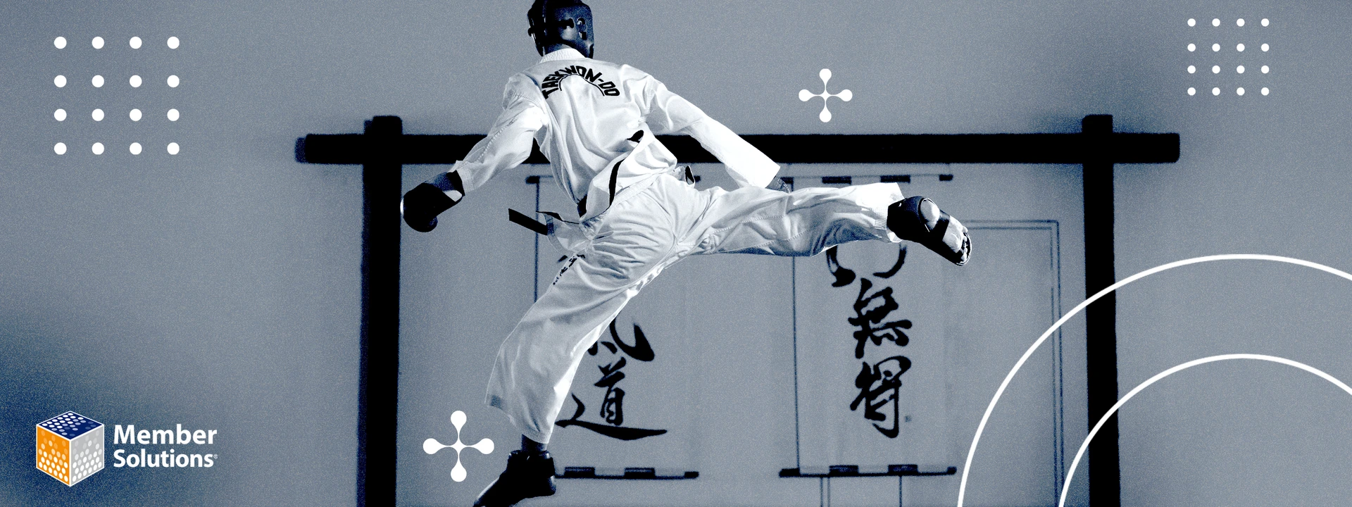 A Taekwondo athlete in a white dobok performs a dynamic flying kick against a wall with Korean calligraphy